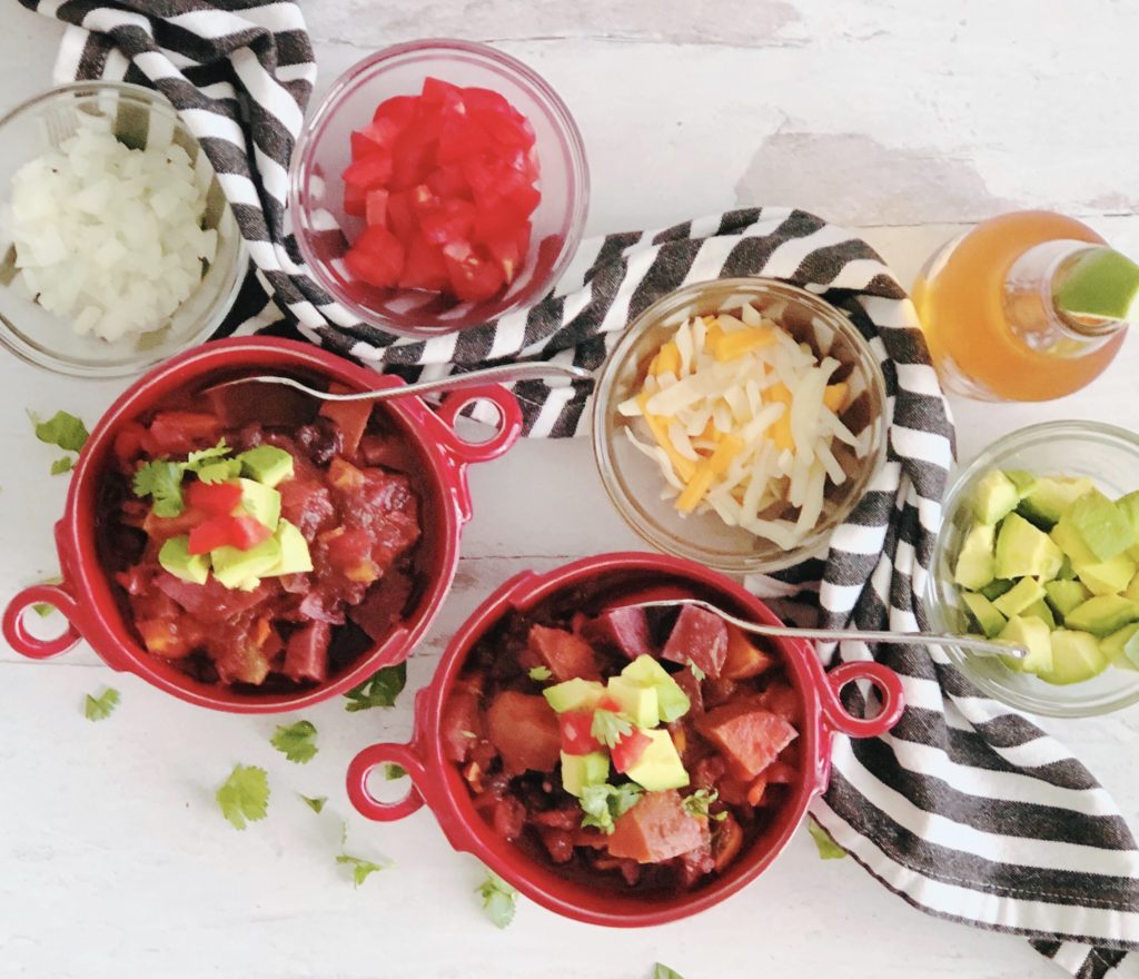 Vegetarian Sweet Potato Chili in red bowls, surrounded by chili toppings in small bowls and a beer.