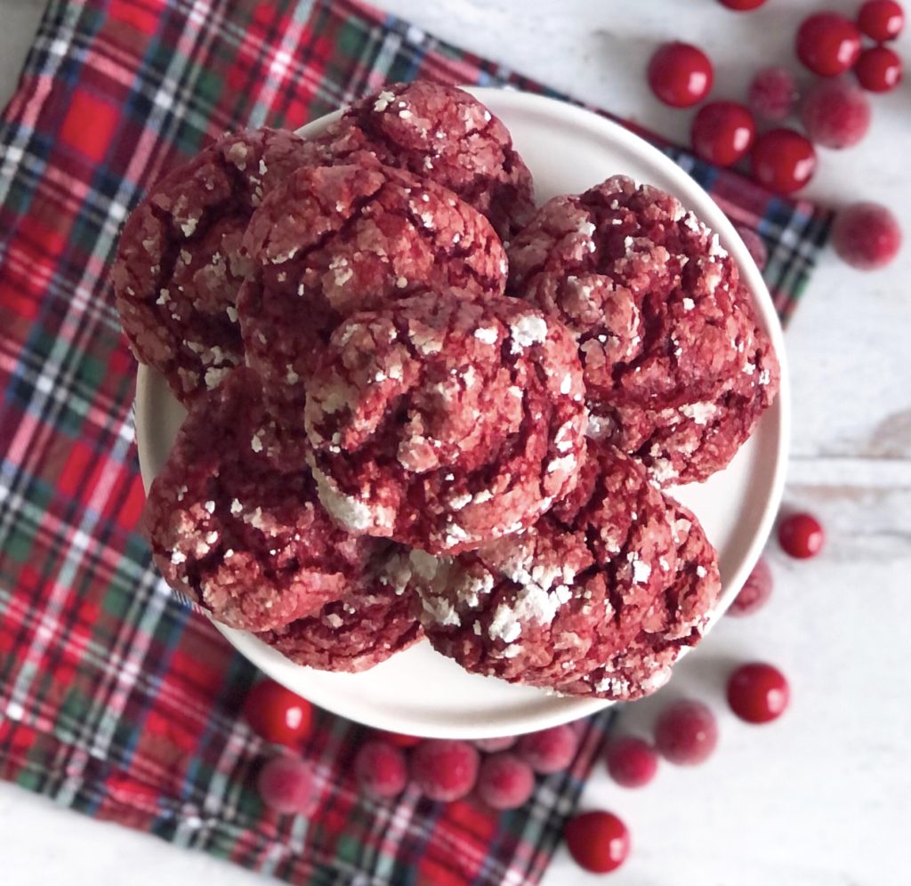 Red Velvet Gooey Butter Cookies on a white pedestal dish sitting on a plaid napkin surrounded by sugared cranberries.