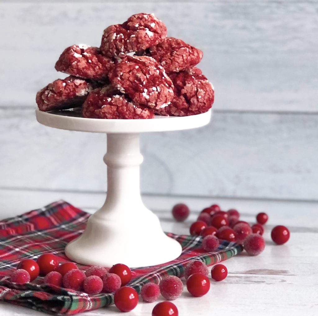 Red Velvet Gooey Butter Cookies on a white pedestal dish sitting on a plaid napkin surrounded by sugared cranberries.