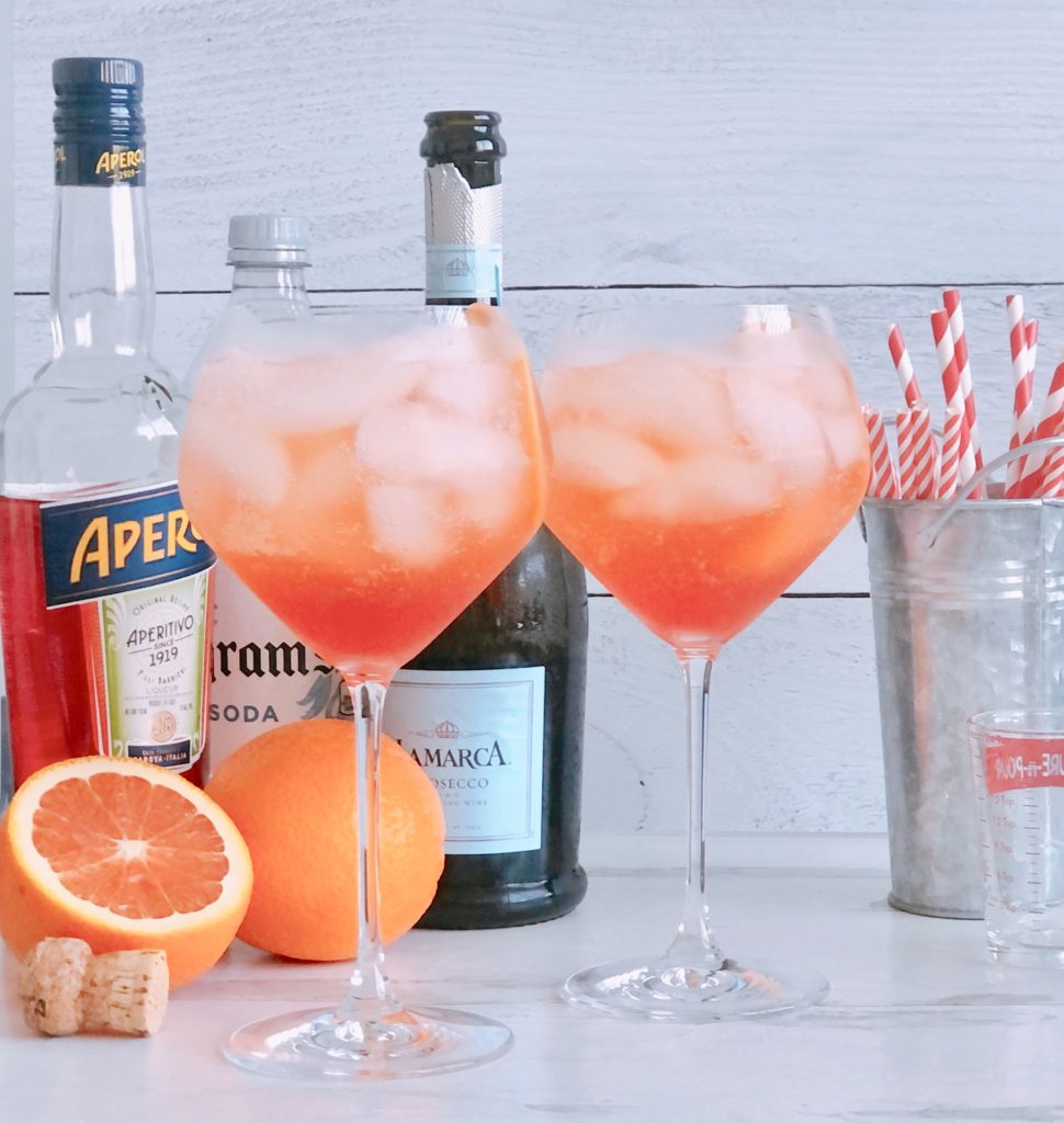 Aperol Spritz sitting in front of Aperol, prosecco, club soda and red and white straws