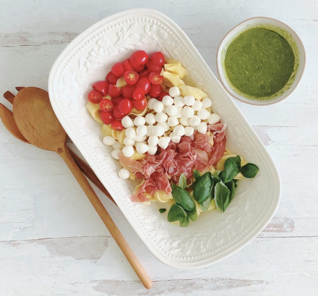 Caprese Pesto Tortellini Salad in a white oval bowl with wooden serving spoons and pesto dressing on the side